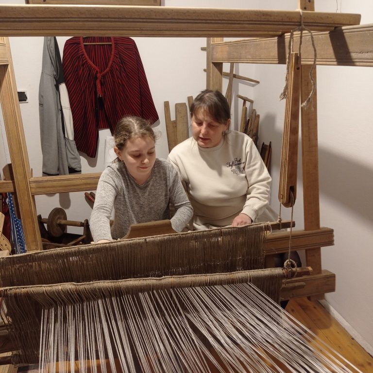 Pani pokazuje dziewczynce jak tkać na krośnie.