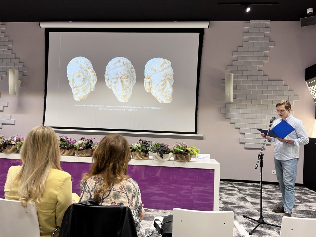 Młody mężczyzna czyta z niebieskiej teczki przy mikrofonie w sali konferencyjnej, na ekranie widoczne trzy rzeźbione głowy z podpisem o kopii przed odtworzeniem warstw malarskich.