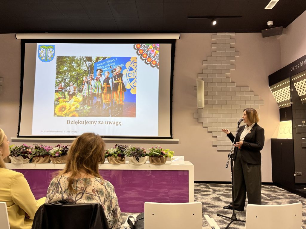 Kobieta w garniturze prowadzi prezentację w sali konferencyjnej, na ekranie widoczny slajd z napisem ‘Dziękujemy za uwagę’ i zdjęciem zespołu folklorystycznego w kolorowych strojach ludowych.