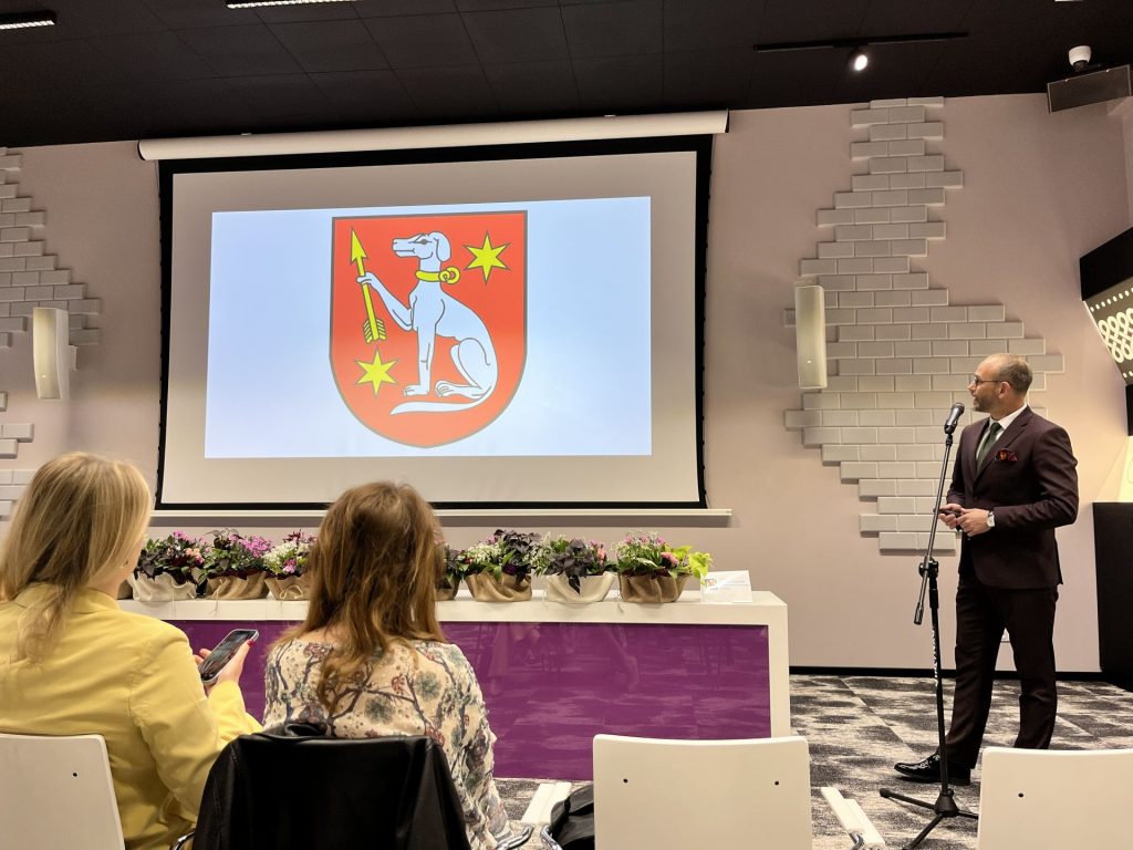 Mężczyzna w garniturze prowadzi prezentację w sali konferencyjnej, na ekranie widoczny herb z białym psem na czerwonym tle.
