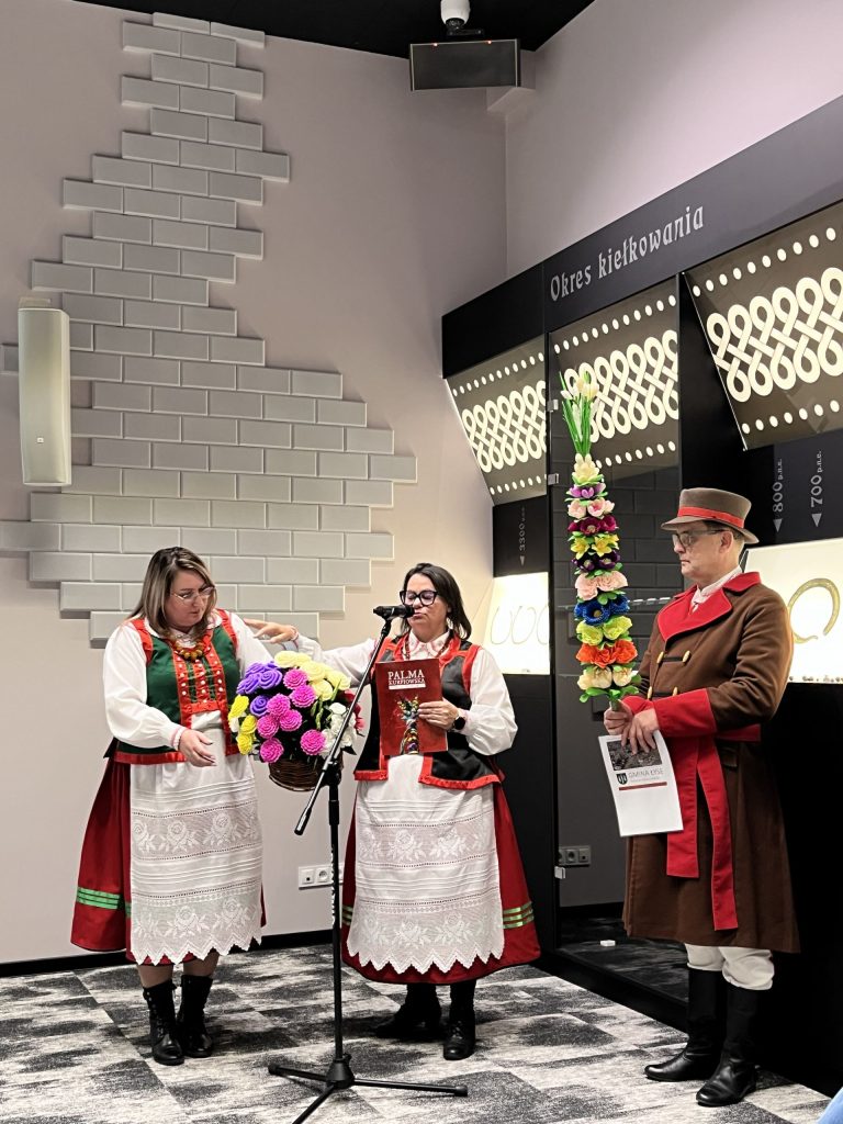 Trzy osoby w strojach ludowych stoją w sali, dwie kobiety prezentują kosz z kolorowymi kwiatami przy mikrofonie, obok mężczyzna trzyma ozdobną palmę wielkanocną.
