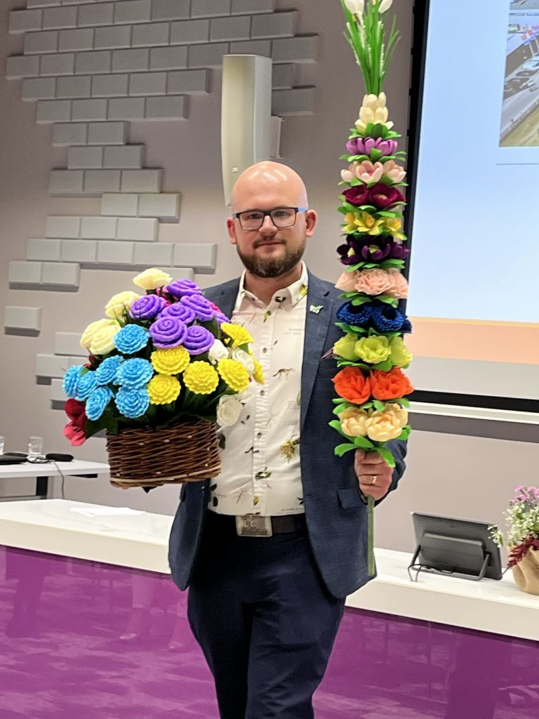 Mężczyzna w garniturze i koszuli w kwiaty prezentuje kosz z kolorowymi kwiatami z bibuły oraz wysoką palmę wielkanocną w sali konferencyjnej.