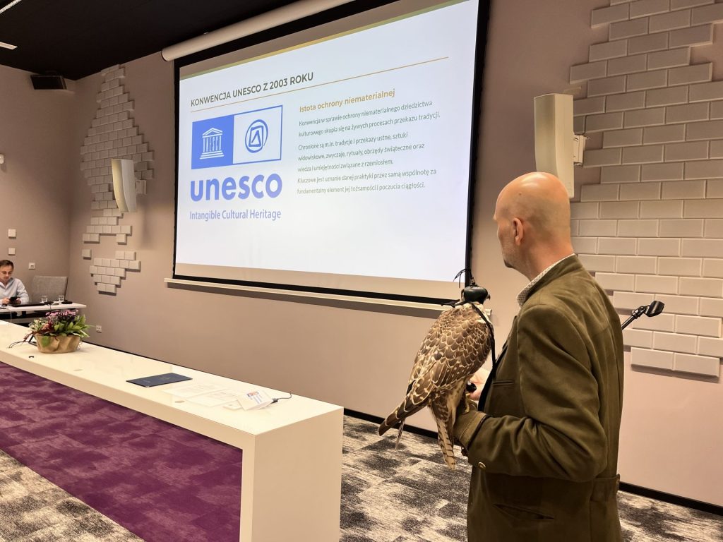 Mężczyzna w brązowej marynarce trzyma sokoła podczas prezentacji o Konwencji UNESCO z 2003 roku, na ekranie widoczny slajd z logo UNESCO i tekstem o ochronie niematerialnego dziedzictwa kulturowego.