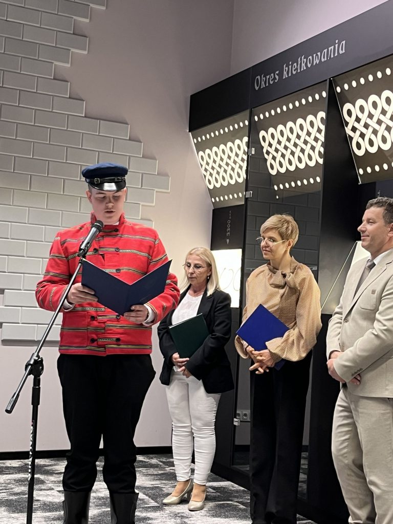 Młody mężczyzna w czerwonym mundurze i czapce czyta z teczki przy mikrofonie, obok stoją trzy osoby z dokumentami.