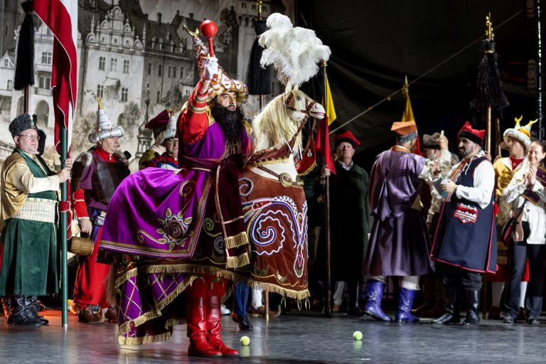Mężczyzna w bogato zdobionym stroju i czerwonych butach, siedzący na stylizowanym koniu scenicznym, unosi berło podczas występu; wokół stoją inni uczestnicy w historycznych kostiumach.