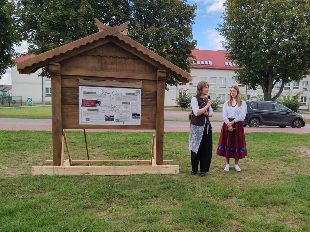 Dwie kobiety stoją obok drewnianej etnotablicy poświęconej miejscowości Złota Góra.
