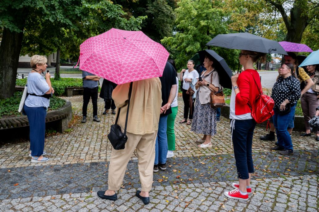 Grupa osób z parasolami słucha przewodniczki podczas spaceru po parku w deszczowy dzień.