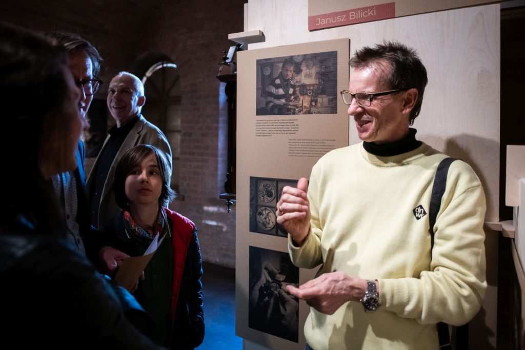 Mężczyzna w żółtym swetrze rozmawia z grupą osób przy wystawie poświęconej Januszowi Bilickiemu w muzeum; w tle ceglane wnętrze i plansza z fotografiami.