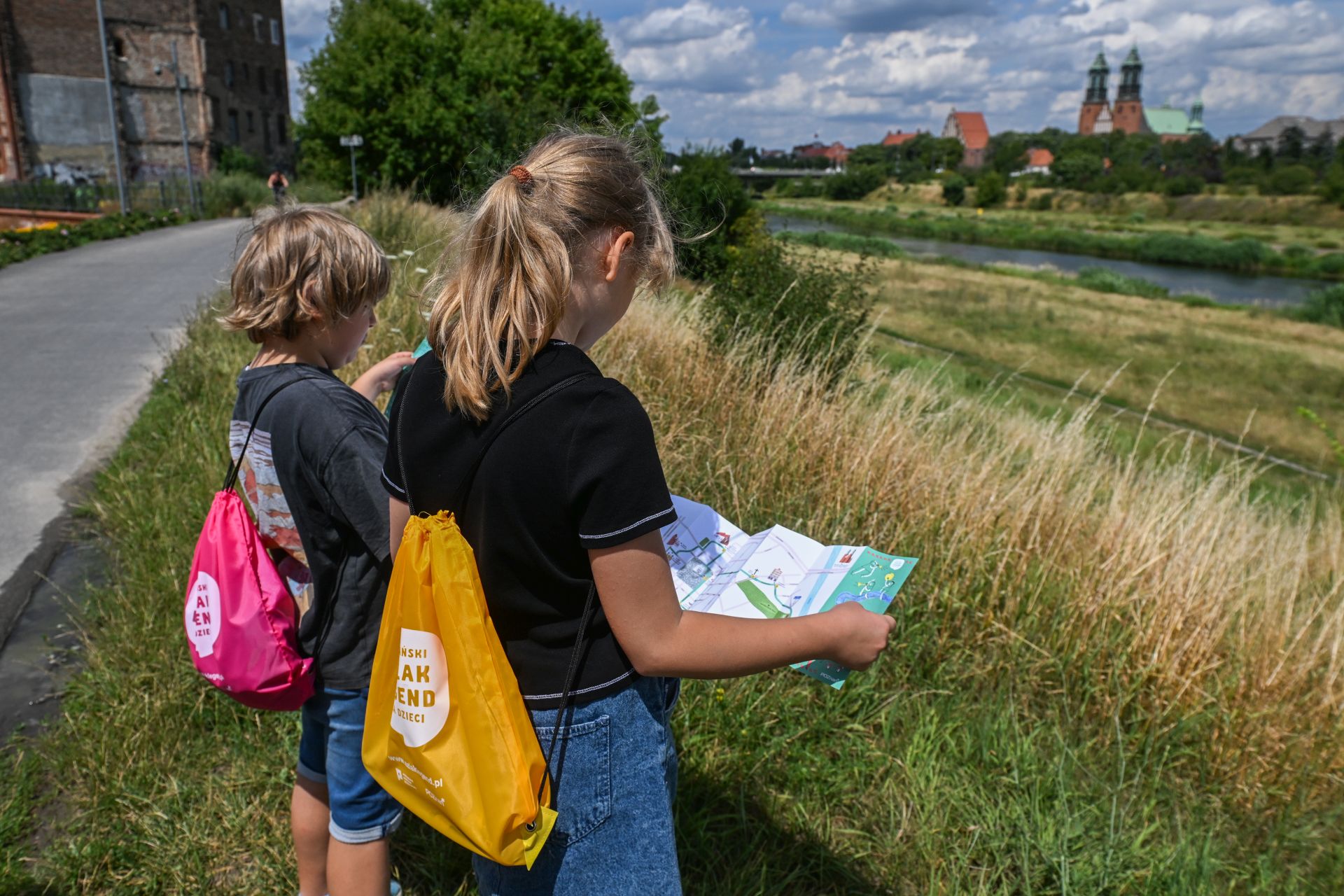 Dwoje dzieci z kolorowymi plecakami ogląda mapę podczas spaceru wzdłuż rzeki, w tle widać katedrę w Poznaniu.