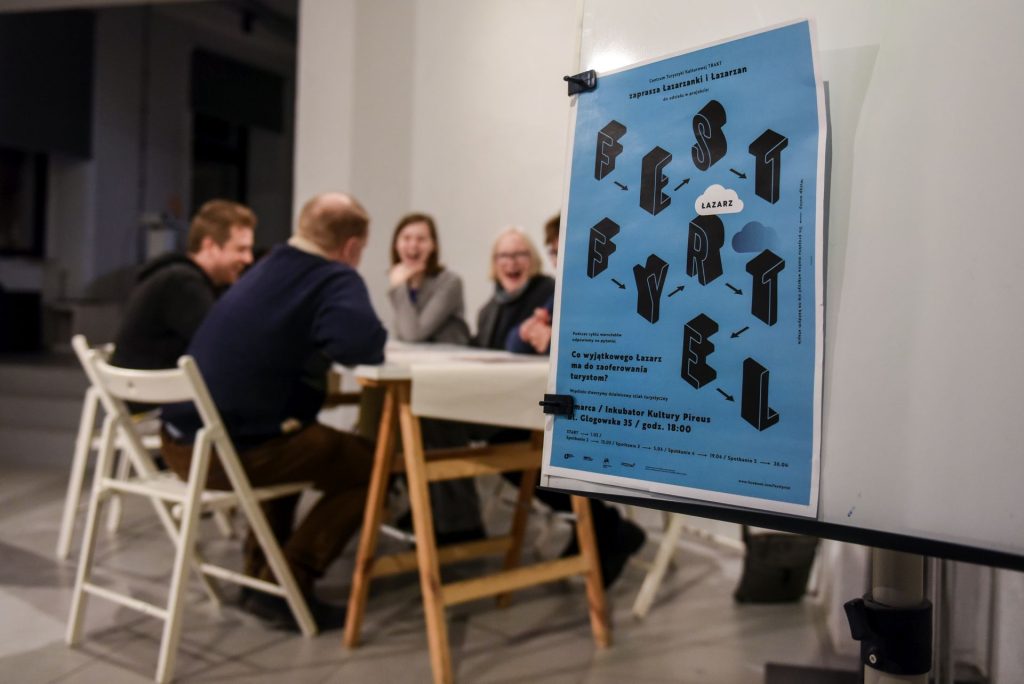 Plakat wydarzenia „Fest Fyrtel Łazarz” na niebieskim tle wisi na stojaku, a w tle grupa osób siedzi przy stole i rozmawia podczas spotkania w jasnym pomieszczeniu.