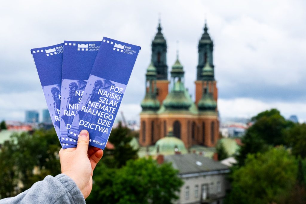 Ręka trzyma trzy niebieskie ulotki „Poznański Szlak Niematerialnego Dziedzictwa” na tle katedry w Poznaniu z zielonymi wieżami i pochmurnym niebem.