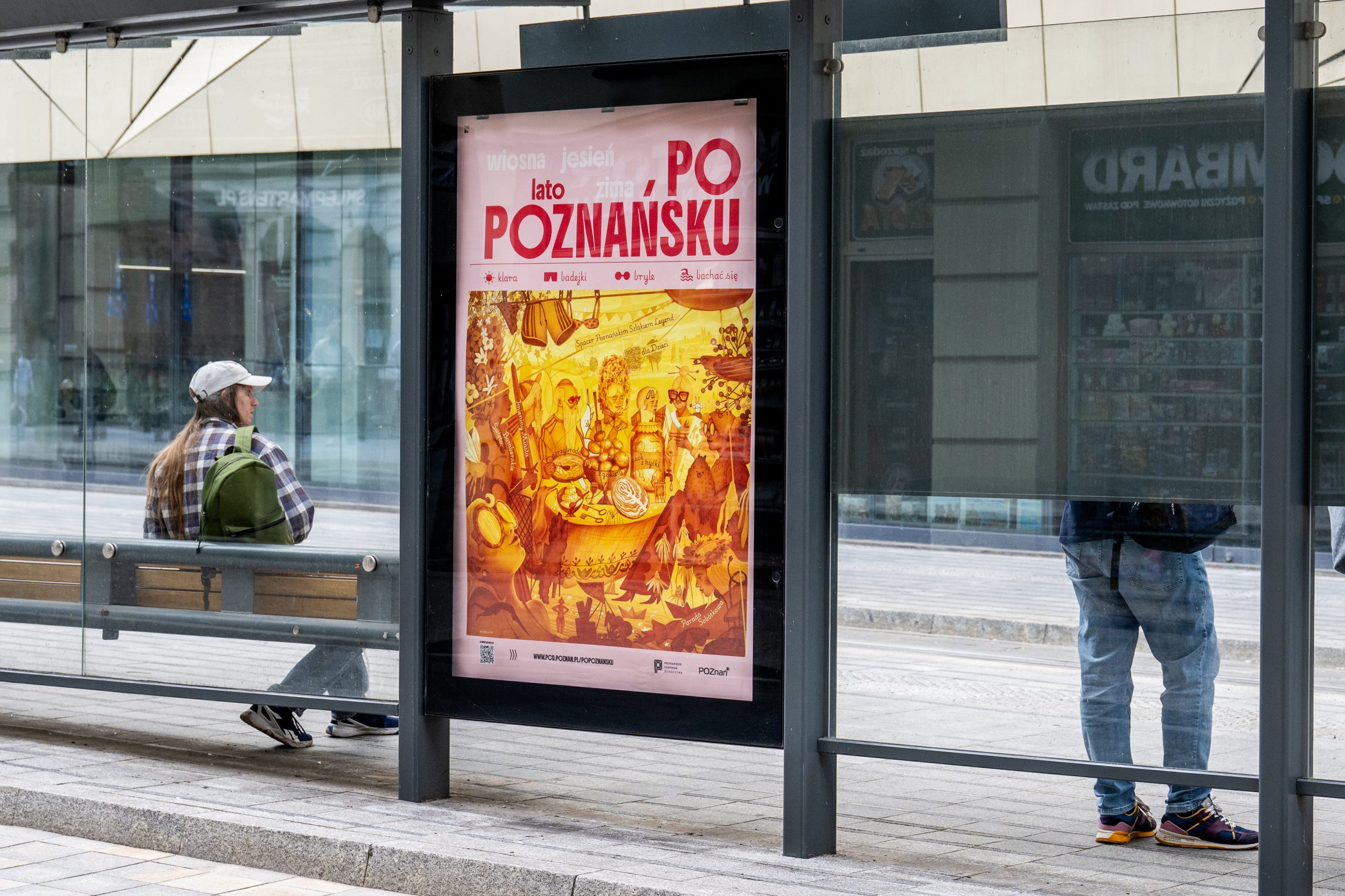 Plakat „Po poznańsku” w witrynie przystanku tramwajowego w centrum miasta; obok siedzi osoba z plecakiem, a w tle widać przechodniów i zabudowę miejską